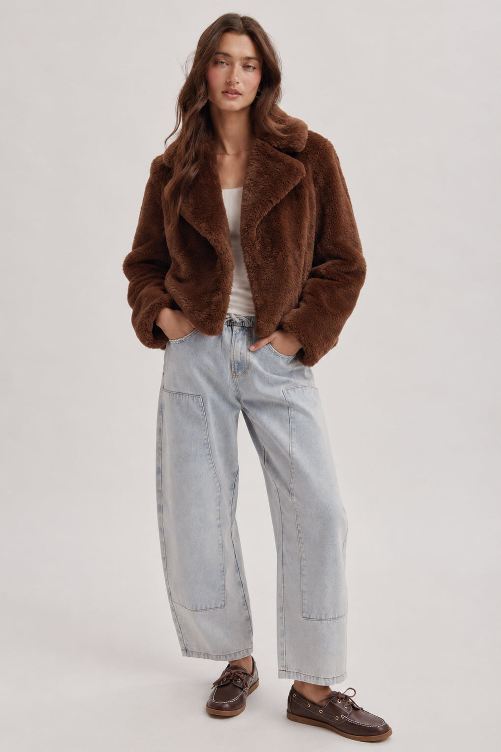 A woman modeling a brown plush faux-fur jacket, paired with light-wash wide-leg jeans, a white top, and brown boat shoes, against a plain light gray studio background.