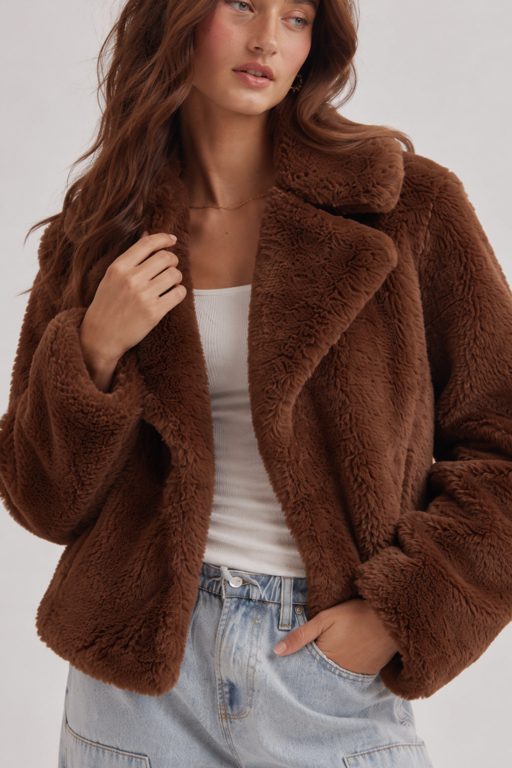 A woman wearing a brown plush teddy faux fur jacket over a white tank top and light wash blue jeans, photographed against a light gray studio background.