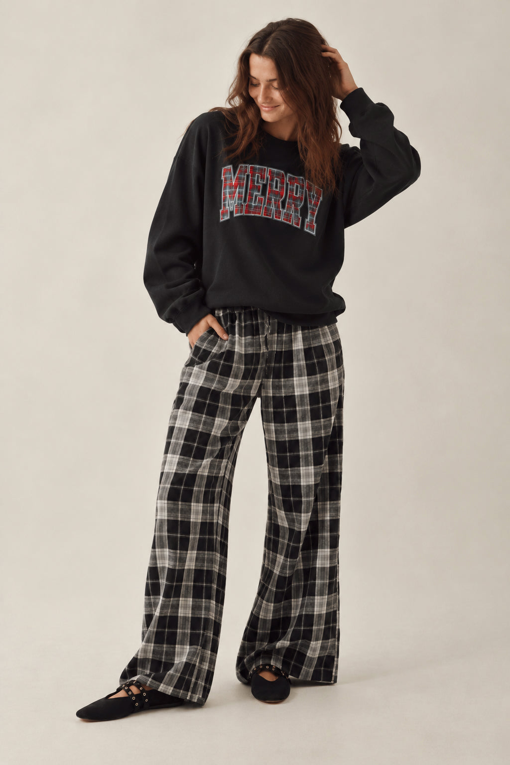 A woman modeling a black sweatshirt with a red and blue plaid 'MERRY' graphic and black-and-white plaid wide-leg pants, standing against a plain light beige studio background.