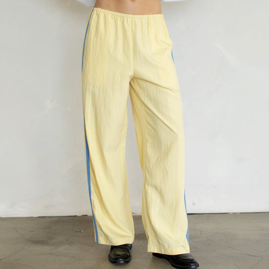 A person wearing wide-leg light yellow pants with blue side stripes, paired with black shoes, standing against a plain off-white wall.