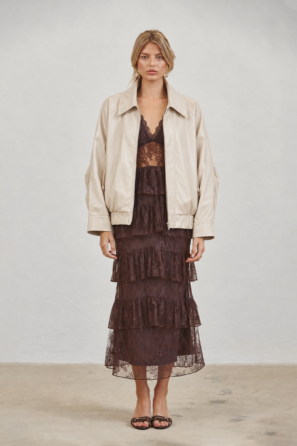 A model stands against a light gray studio wall wearing a cream-colored leather jacket over a dark brown lace tiered dress and black flat sandals.