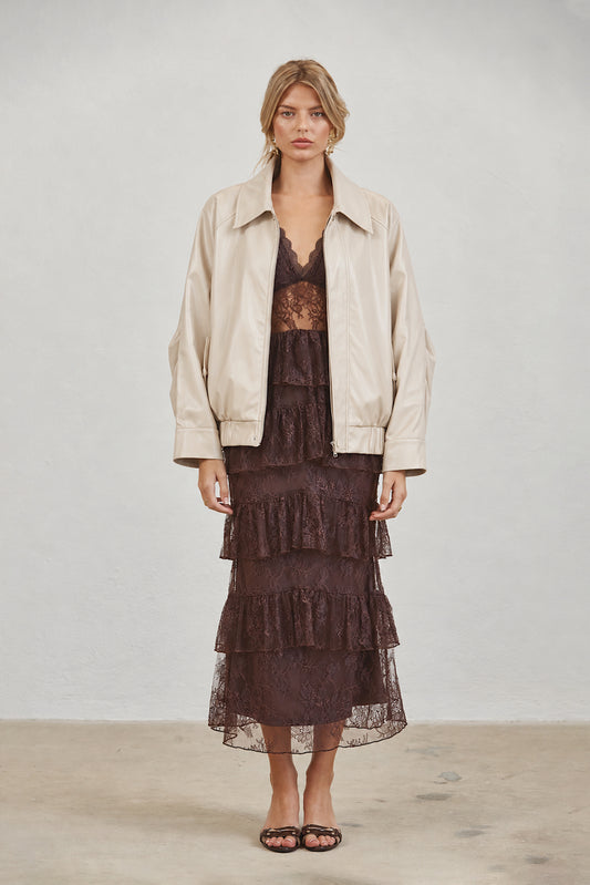 A model stands against a light gray studio wall wearing a cream-colored leather jacket over a dark brown lace tiered dress and black flat sandals.