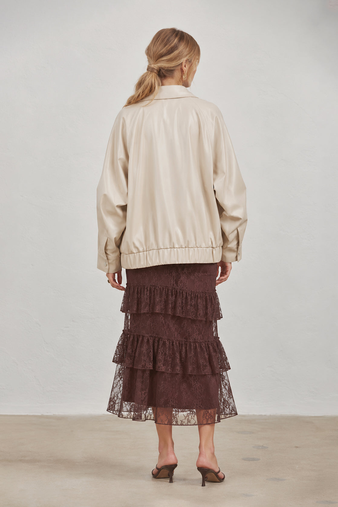 A woman shown from the back wearing a cream faux-leather bomber jacket and a dark brown tiered lace midi skirt, standing against a neutral light gray studio background.