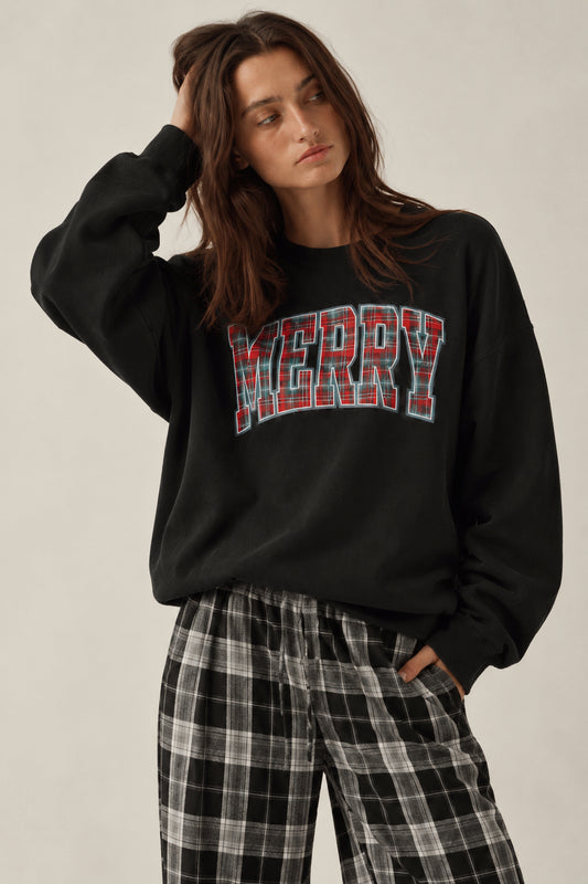 A woman wearing a black crewneck sweatshirt with 'MERRY' printed across the chest in red and white plaid letters, paired with black and white plaid pajama pants, against a plain light beige background.