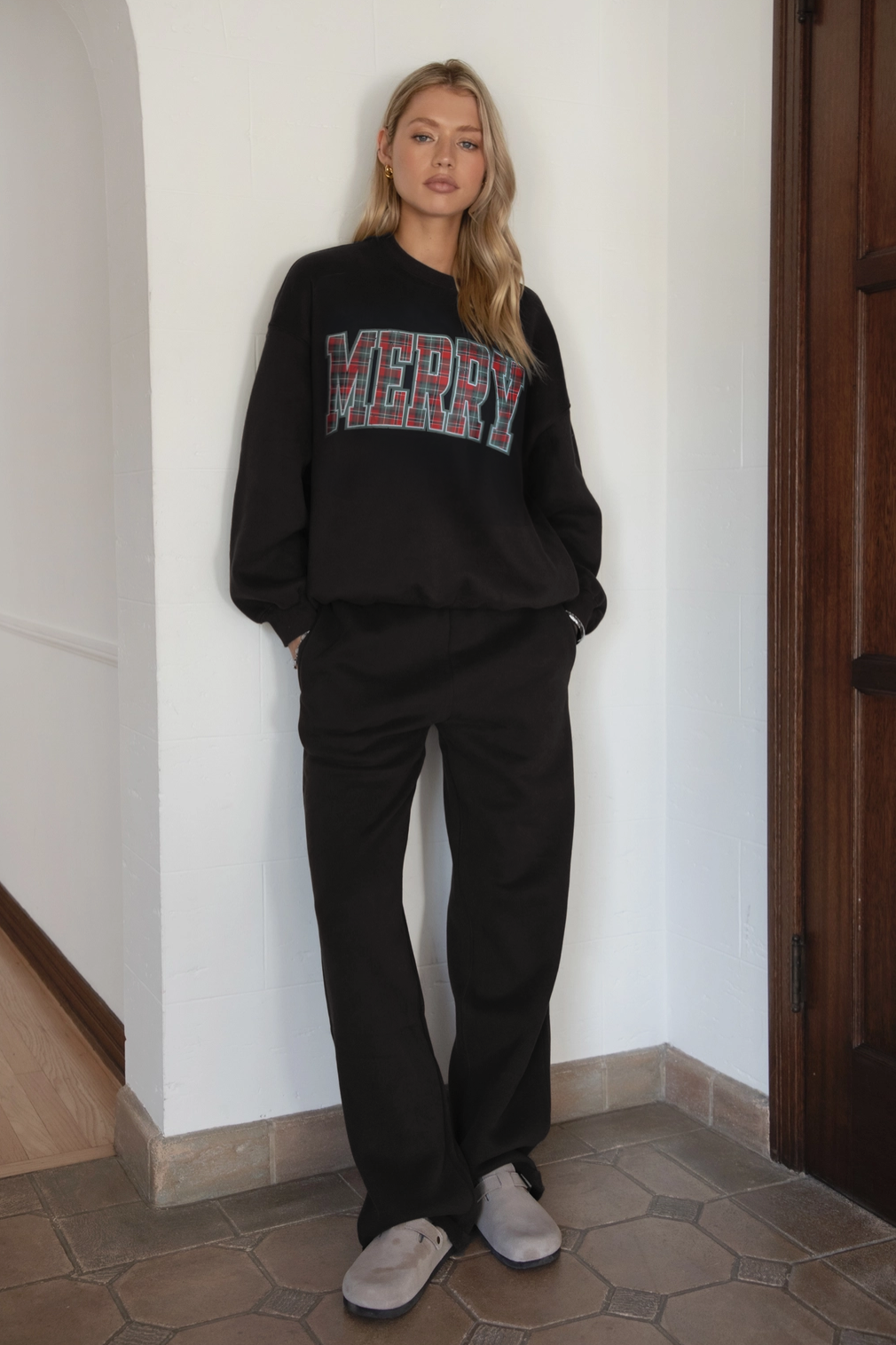 Person wearing a black sweatshirt with 'MERRY' text, standing against a white wall.