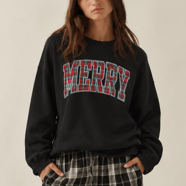 A woman wearing a black crewneck sweatshirt with 'MERRY' printed across the chest in red and white plaid letters, paired with black and white plaid pants, against a plain beige background.