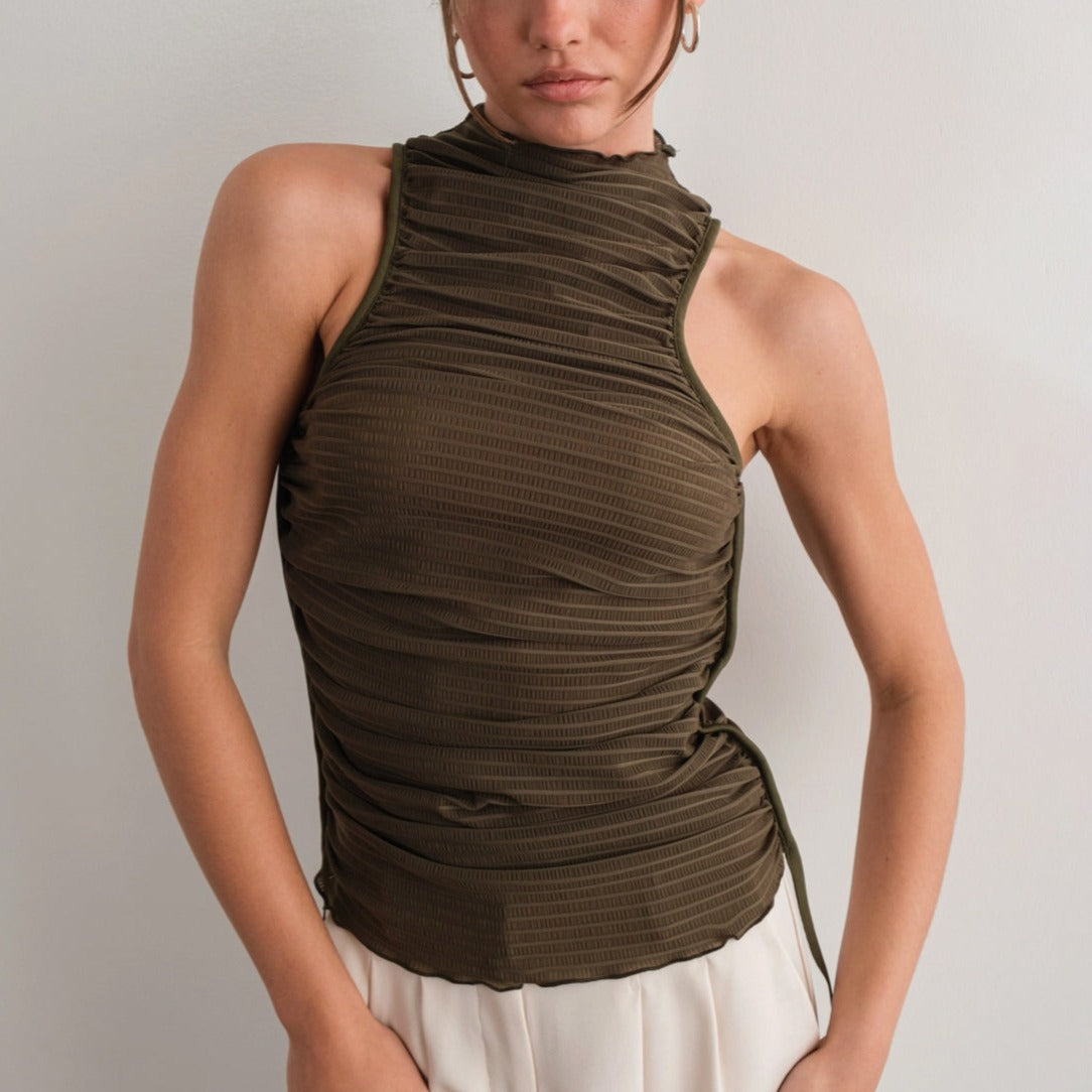 A person wearing an olive green high neck ruched tank top with a lettuce-edge mock neck and low-cut armholes, paired with a white skirt.