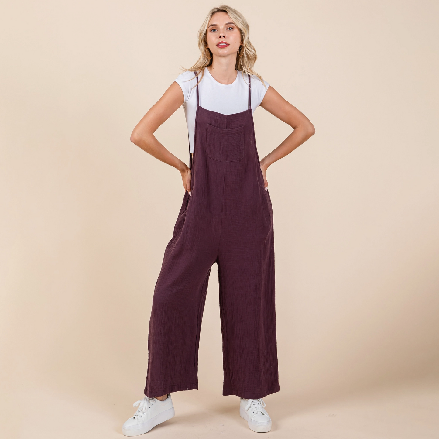 A female model standing against a beige background, wearing a white t-shirt underneath a terracotta-colored overall jumpsuit with wide-leg and adjustable straps.