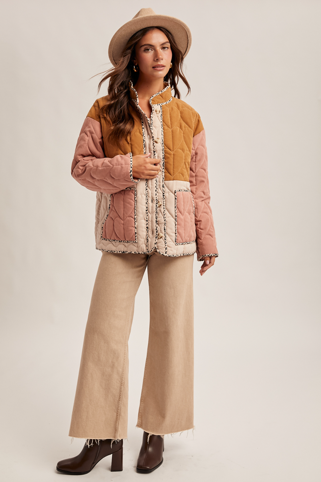 Woman wearing a colorful quilted jacket, beige pants, and a hat on a plain background