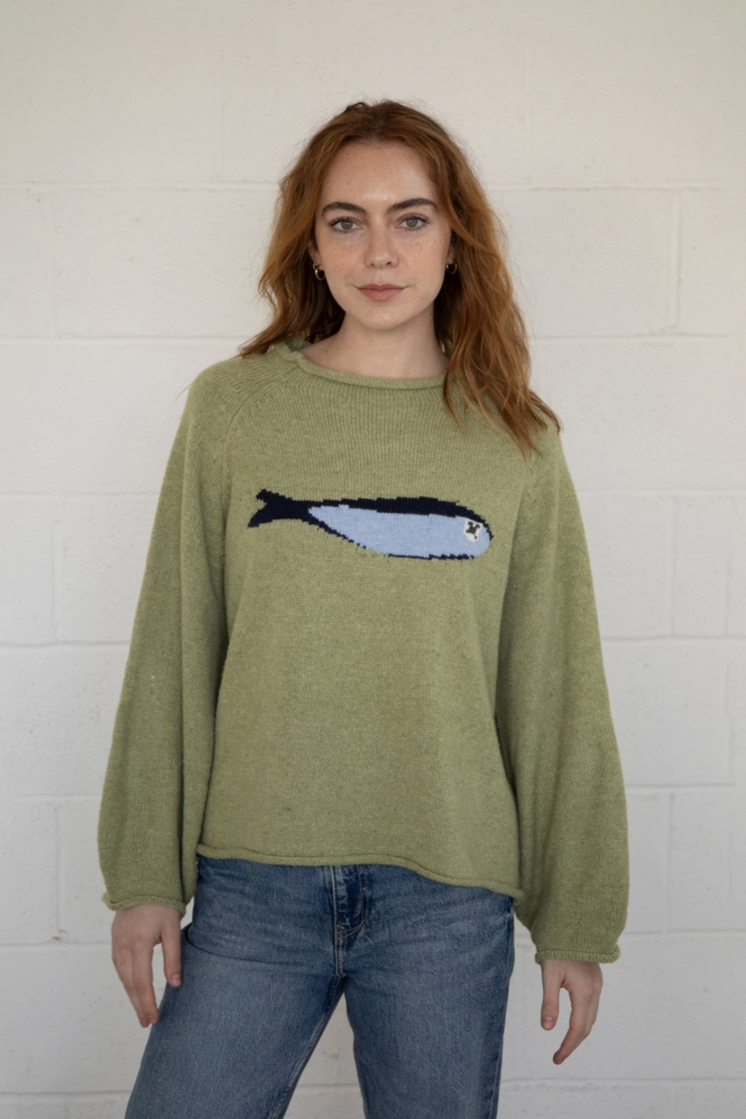 Woman wearing a green sweater with a fish design against a white brick wall.