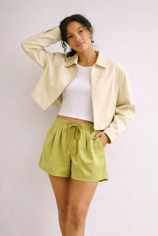 Woman wearing a light yellow jacket, white top, and green shorts against a plain background