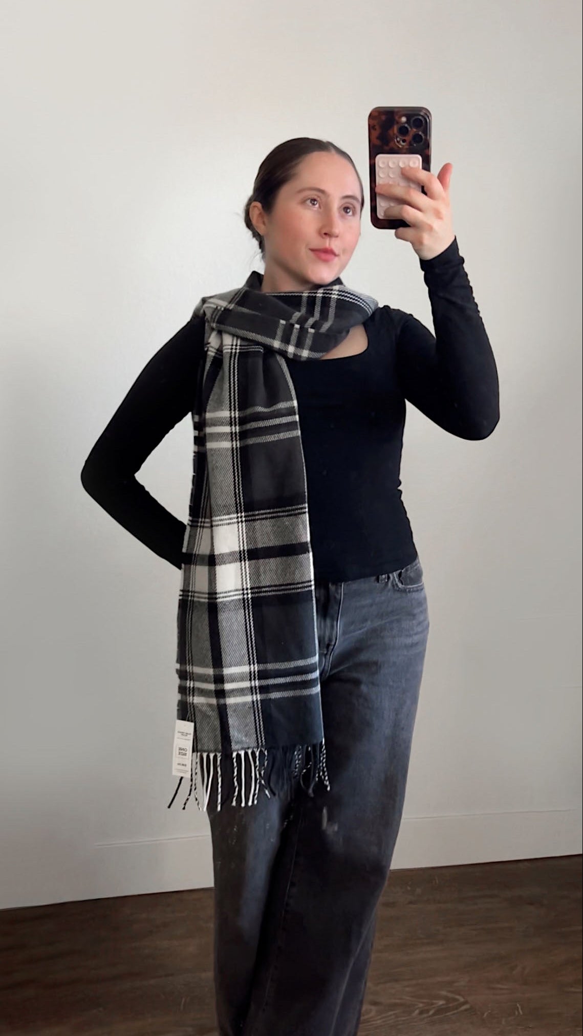 Person wearing a plaid scarf, black shirt, and jeans taking a mirror selfie.