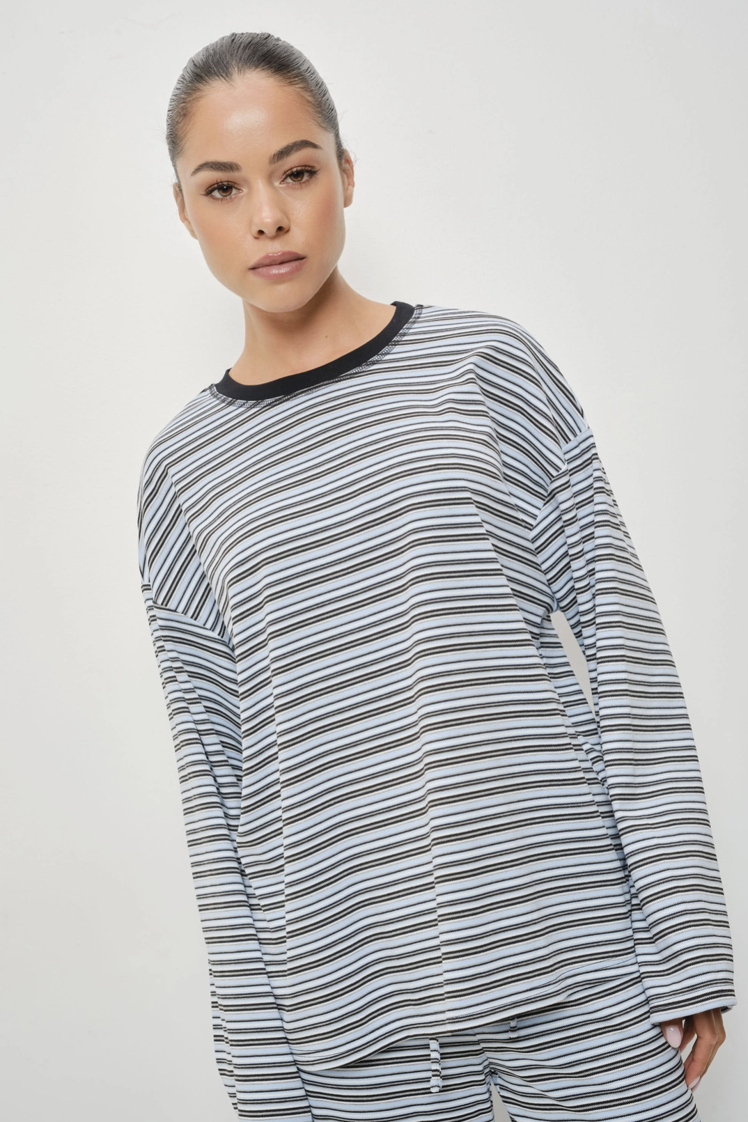 A woman wearing a long-sleeve pajama top with thin horizontal black, gray, and white stripes, featuring a solid black crew neck, photographed against a light gray studio background.