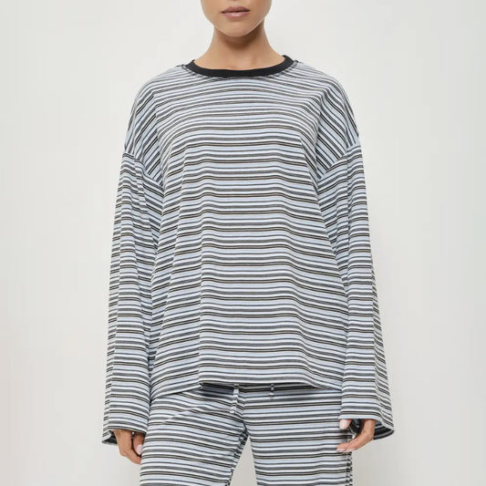 A model wearing a matching long-sleeve striped lounge set, featuring a loose crewneck top and coordinating pants in a gray, black, and white horizontal stripe pattern.
