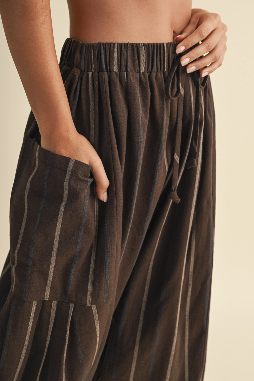 Close-up of a person wearing loose brown drawstring pants with vertical light stripes and a side pocket, against a warm beige background.