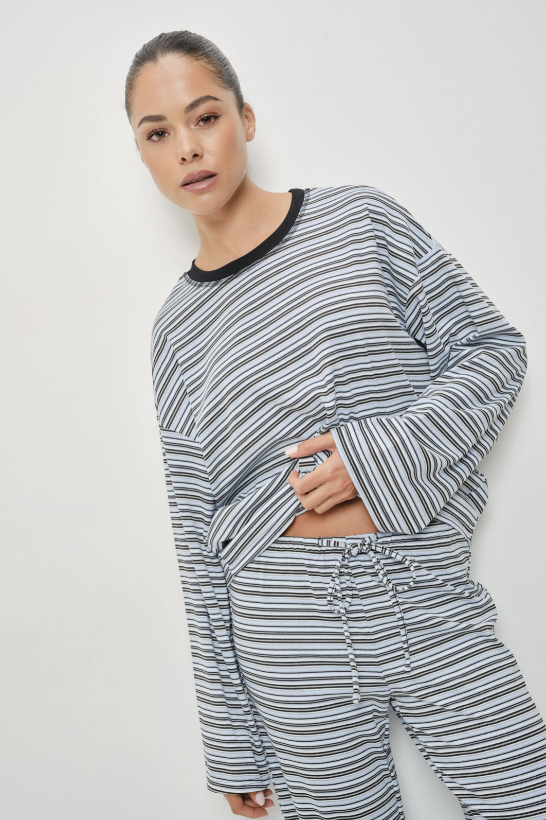 A woman modeling a matching long-sleeve striped lounge set with a black crew neck and drawstring waist, photographed against a plain light gray studio background.