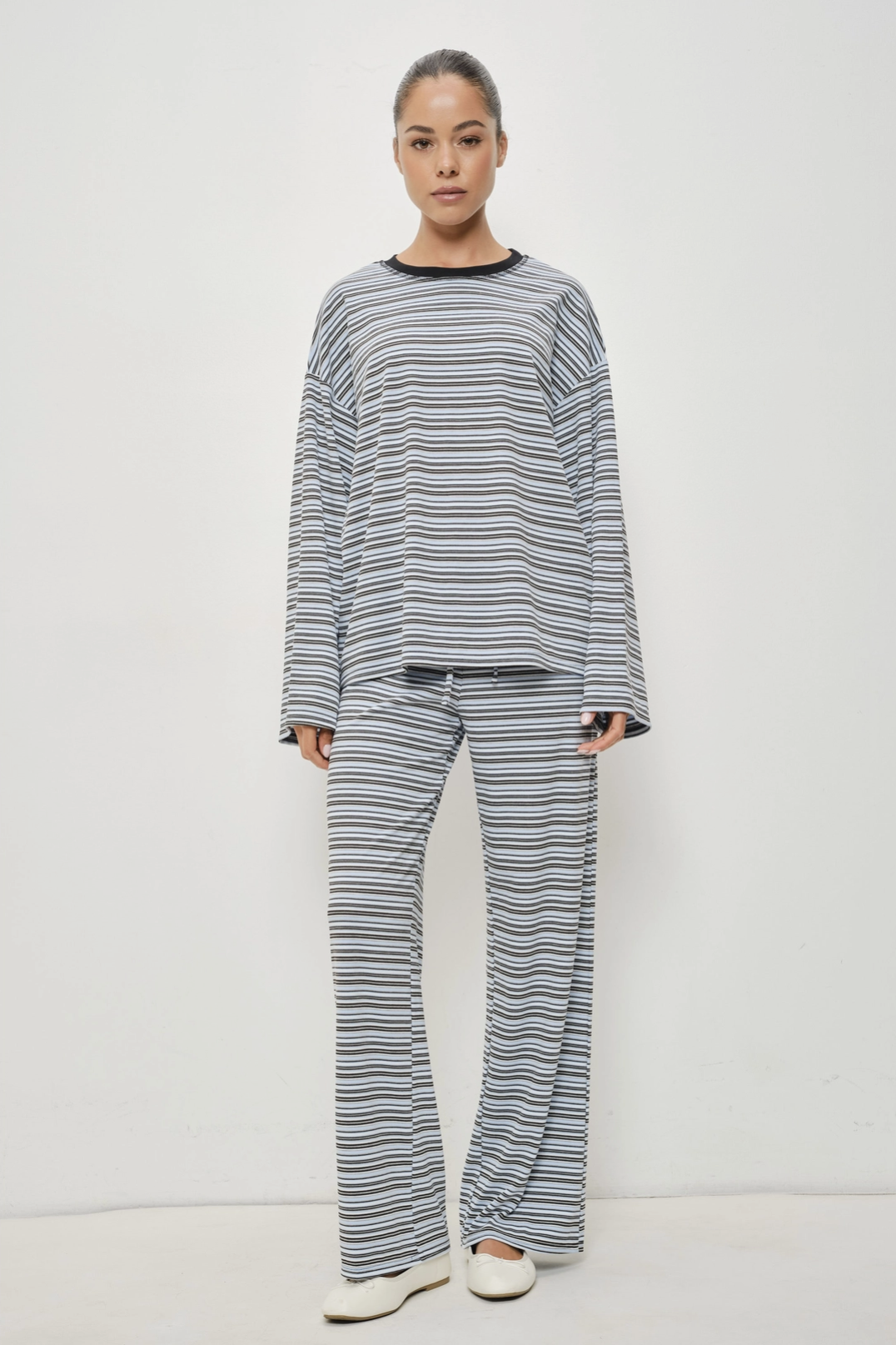 A woman modeling a matching two-piece loungewear set consisting of a long-sleeve top and wide-leg pants, both featuring a fine horizontal stripe pattern in gray and white with a black crew neck trim.