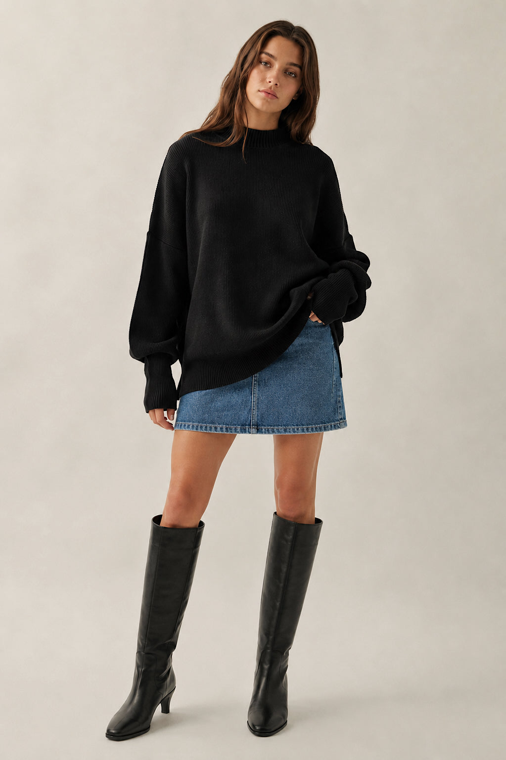 Woman wearing a black sweater, blue denim skirt, and black knee-high boots on a plain background