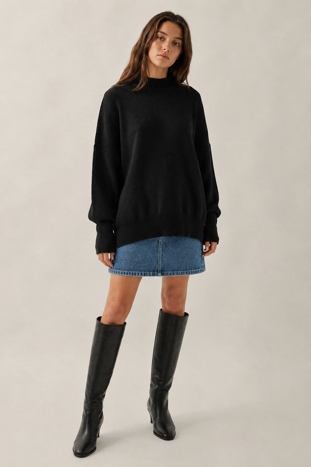 Woman wearing a black sweater, blue denim skirt, and black knee-high boots on a plain background