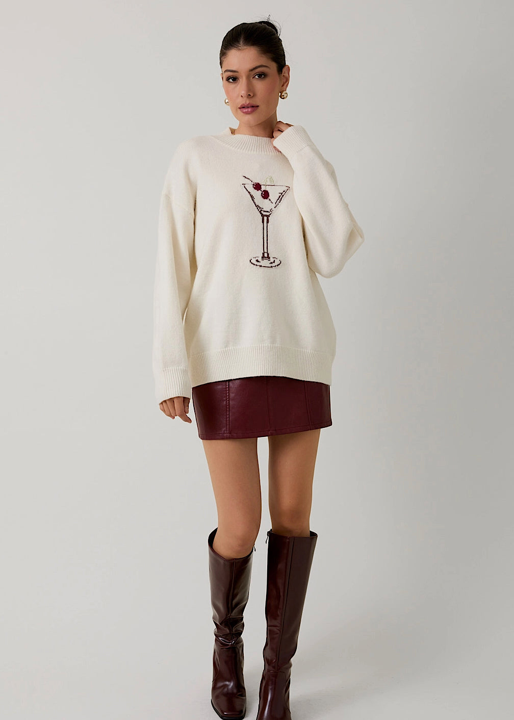 Woman wearing a cream sweater with a martini glass design, burgundy leather skirt, and brown knee-high boots on a white background.