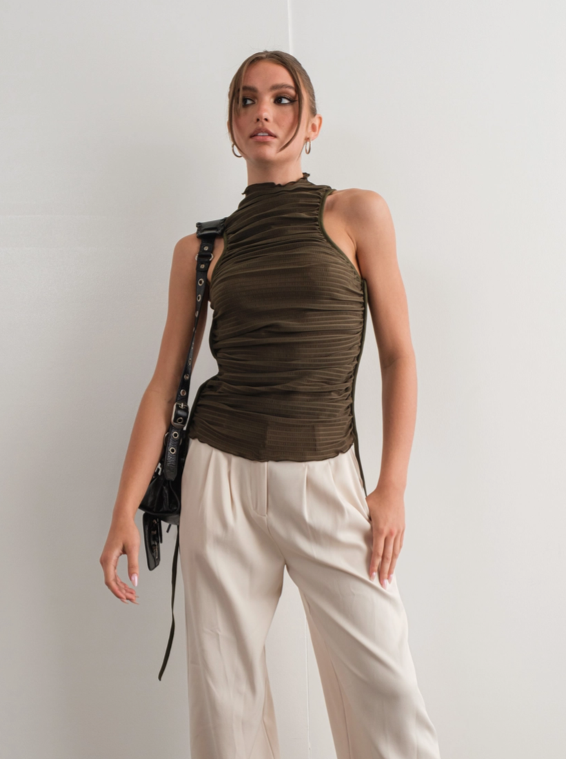 A person wearing an olive green high neck ruched tank top with a lettuce-edge mock neck and low-cut armholes, paired with a white skirt.