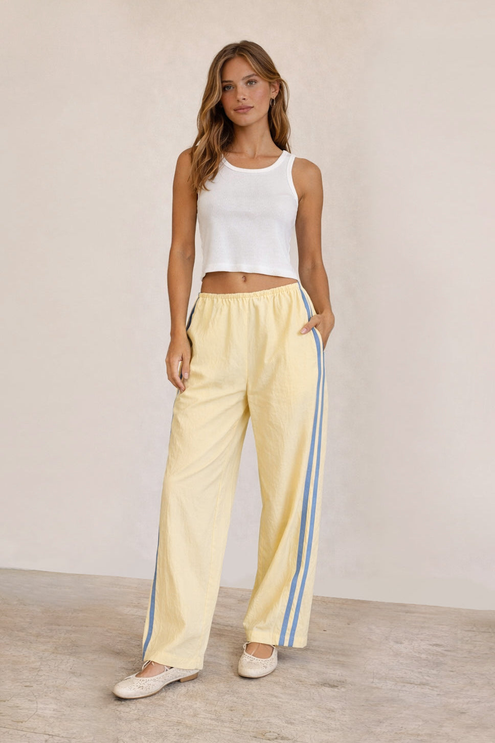 Woman wearing a white sleeveless top and light yellow pants with blue stripes on a plain background